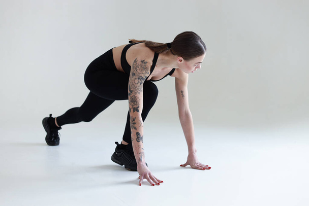 A young girl with brown hair, with a tattoo in a black top and leggings on a white background. Sports, fitness, starts running - Photo, Image
