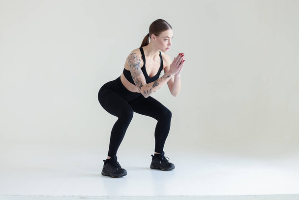 A young girl with brown hair, with a tattoo in a black top and leggings on a white background. Sports, fitness, squats - Φωτογραφία, εικόνα
