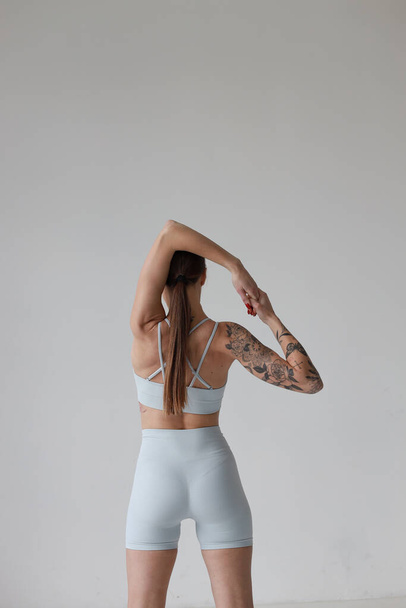 Jong meisje met bruin haar, met een tatoeage in een grijs topje en korte broek op een witte achtergrond met hoofdtelefoon. Sport, fitness, stretchen en yoga, meditatie - Foto, afbeelding