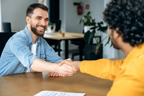 The concept of a deal, agreement. Two men of different nationalities, sitting at a desk in office, shaking hands after successful negotiations or job interview, agreed on cooperation, signed contract - Foto, afbeelding
