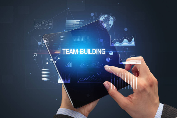 Businessman holding a foldable smartphone with TEAM BUILDING inscription, successful business concept - Photo, image