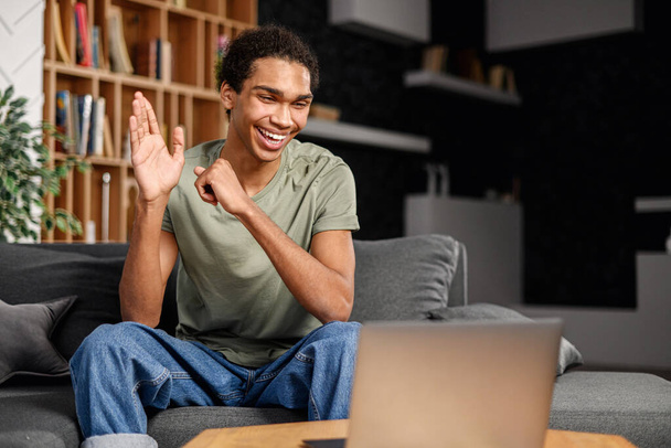 Cheerful multiracial man in casual wear is sitting on the comfortable sofa and using trendy laptop for video call, waving hand, greeting online interlocutor. Virtual meeting, video call concept - Фото, зображення