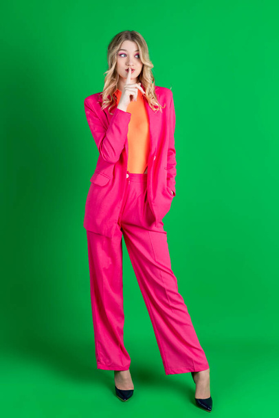 Young curly blonde dressed in pink blazer and pants, gesturing quiet sign with a hand. Isolated on green background. - Photo, image