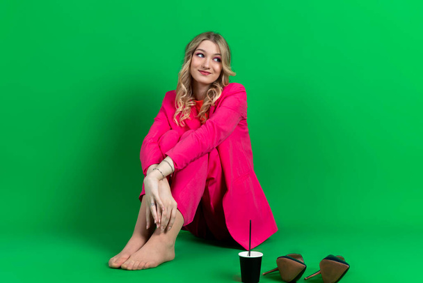 Молода щаслива кучерява блондинка, одягнена в рожевий блейзер і штани, сидить з взуттям
 - Фото, зображення