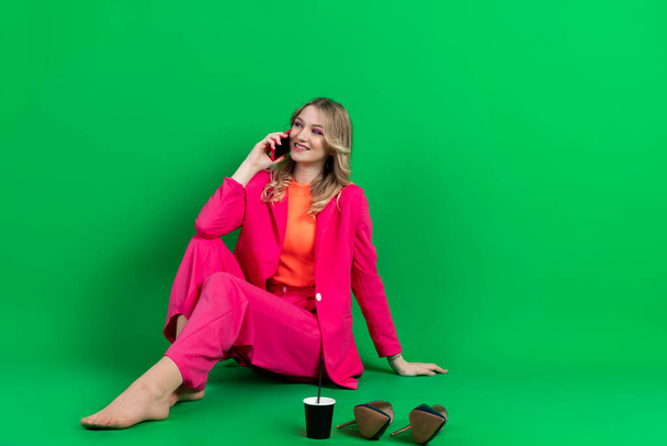 Young happy curly blonde dressed in pink blazer and pants, sitting with shoes off, talking on a phone, calling - Foto, afbeelding