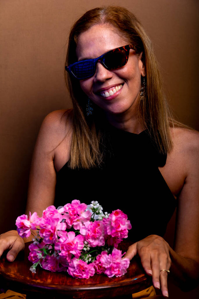 Lachende, gelukkige vrouw zittend op een houten kruk met een zonnebril, met een boeket bloemen. Geïsoleerd op bruine achtergrond. - Foto, afbeelding