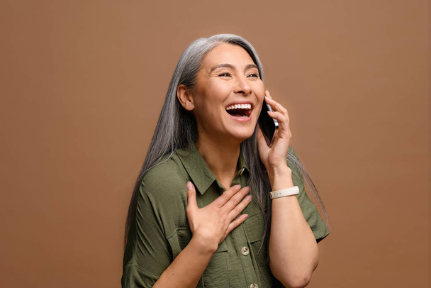 Friendly hilarious mature asian woman talking casually with friend via smartphone, satisfied good mobile connection gray-haired lady holding phone call, enjoying chatting by phone, laughing isolated - Foto, Imagem