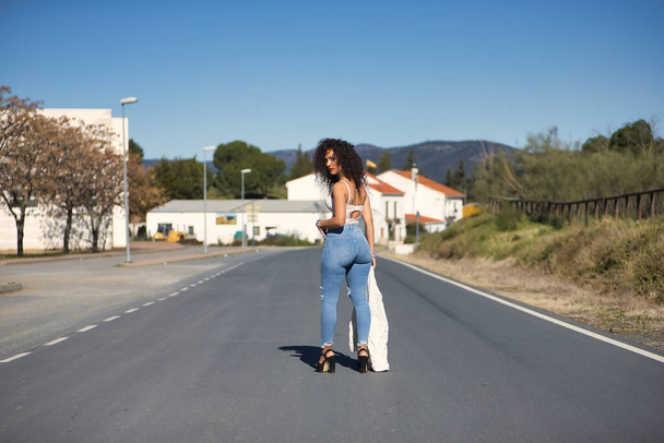 Young, pretty, brunette woman with curly hair, white top, jeans and heels, standing posing in the middle of a lonely road. Concept beauty, fashion, posing, model. - Valokuva, kuva