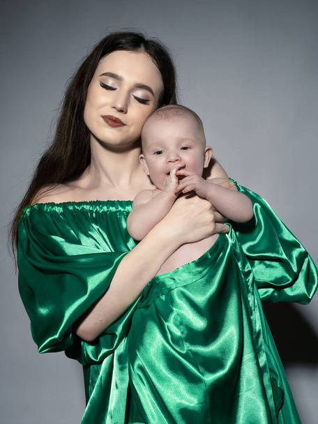 A young, modern, beautiful loving mommy holds hands, kisses her adorable baby in a bodysuit, expresses affection, feels love, close-up. Happy mother's day, motherhood, carefree childhood, guardianship concept - Photo, Image