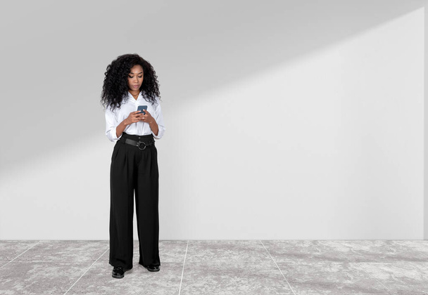 Black businesswoman typing in phone in hands, full length on tile floor. Concept of online communication and social media. Copy space empty wall - Fotoğraf, Görsel