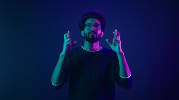 Reményteli indiai férfi muszlim férfi modell szemüvegben Arab srác ujjak keresztbe imádkozni kívánság győzelem győzelem szerencse imádkozó gesztus remény a jó eredményeket kívánság sötét kék neon stúdió háttér - Felvétel, videó