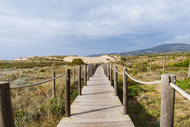 Порожня, дерев'яна прогулянка на пляжі Praia do Guincho в Сінтрі. Погляд на траву й пісок з пагорбами й Атлантичним океаном на задньому плані. - Фото, зображення