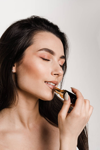 Attractive model is applying matte pastel lipstick on her lips. Girl make-up artist paints her lips with lipstick on white background - Φωτογραφία, εικόνα