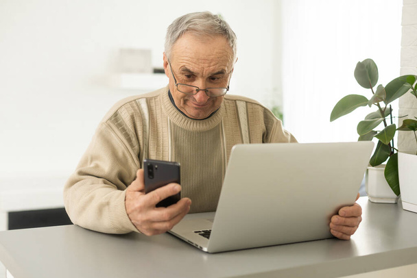 Concentrated senior man looking at laptop and phone calling at home - 写真・画像