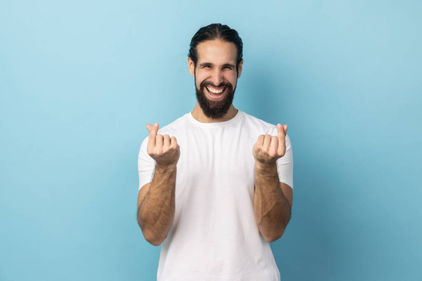 Portrait of smiling happy positive man with beard wearing white T-shirt hair makes korean like sign mini hear gesture snaps fingers. Indoor studio shot isolated on blue background. - Fotografie, Obrázek