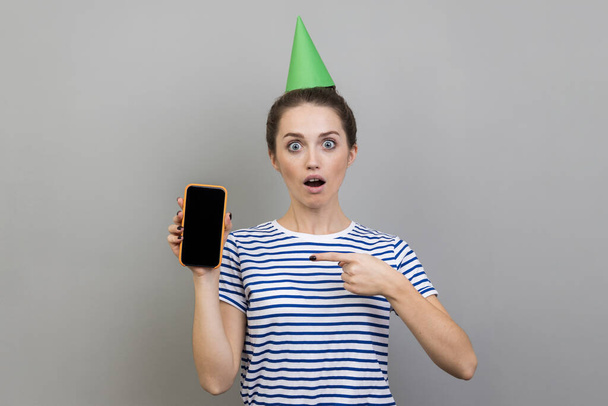 Portrait of surprised astonished woman wearing striped T-shirt and party cone, showing pointing mobile phone with empty black screen for advertisement. Indoor studio shot isolated on gray background. - Fotoğraf, Görsel