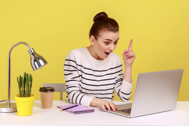 Amazed woman manager in striped shirt looking surprised by genius idea, raising finger up inspired while working on laptop. Indoor studio studio shot isolated on yellow background. - Photo, image