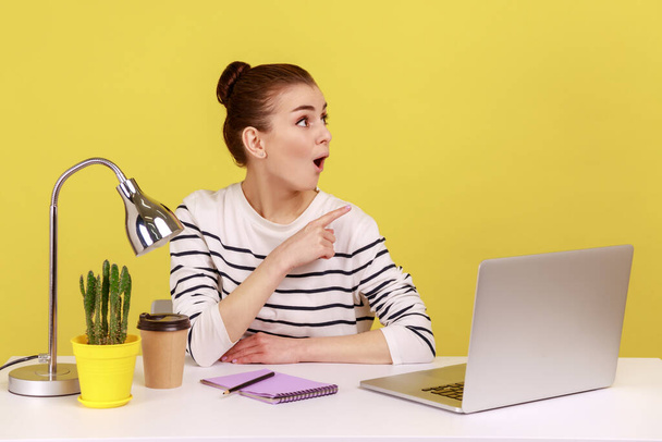 Shocked amazed young woman sitting at office workplace with laptop, looking and pointing aside, showing copy space for promotional text. Indoor studio studio shot isolated on yellow background - Φωτογραφία, εικόνα