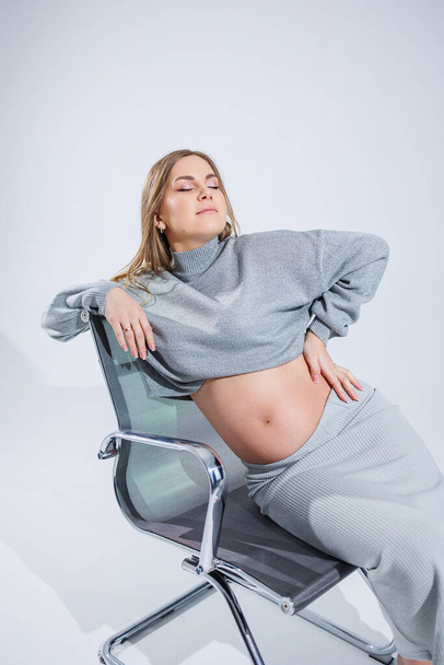 Cute young pregnant woman sitting on a chair on a white background. Stylish pregnant woman in skirt posing in studio and enjoying pregnancy - Fotó, kép