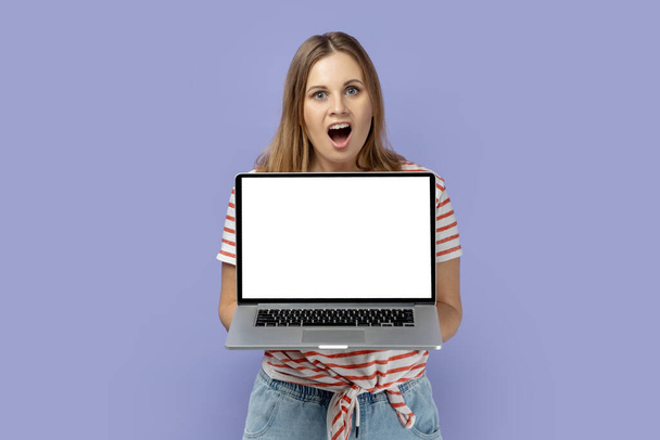 Portrait of shocked amazed blond woman wearing striped T-shirt holding laptop with white empty display with empty space for advertisement. Indoor studio shot isolated on purple background. - Foto, Bild