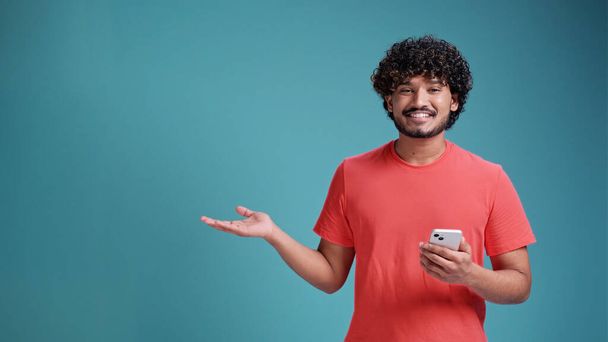 Young indian man using smartphone standing very happy pointing with hand and finger to the side, in coral t-shirt on blue studio background. - Фото, зображення