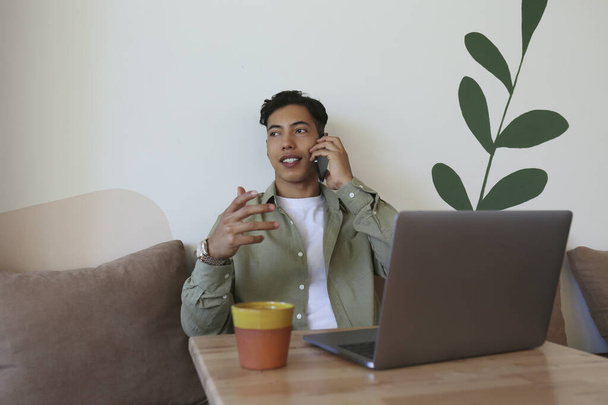 Hispanic young man sitting in coffee shop with his laptop and working. Latino freelancer having a phone call. Remote work concept. Close up, copy space, background. - Foto, afbeelding