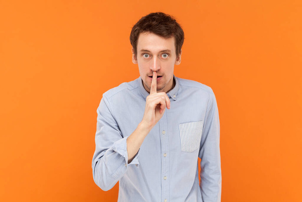 Serious young adult man standing with finger near lips, asking to keep silent, looking at camera with bossy expression, wearing light blue shirt. Indoor studio shot isolated on orange background. - Photo, Image