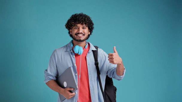 indian or spanish male student with backpack and laptop showing thumbs up gesture of approval while standing against blue studio background. - Foto, Imagem