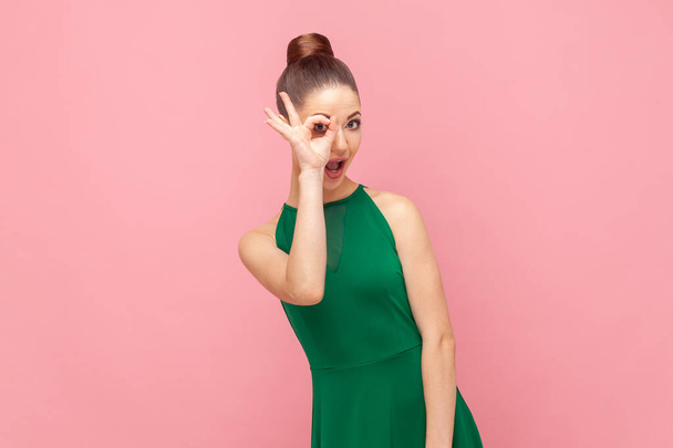 Portrait of funny childish young adult woman standing covering her eye with ok gesture, looking at camera, showing okay sign, wearing green dress. Indoor studio shot isolated on pink background. - Photo, image