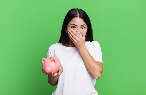 hispanic pretty woman covering mouth with hands with a shocked with a piggy bank - Fotoğraf, Görsel