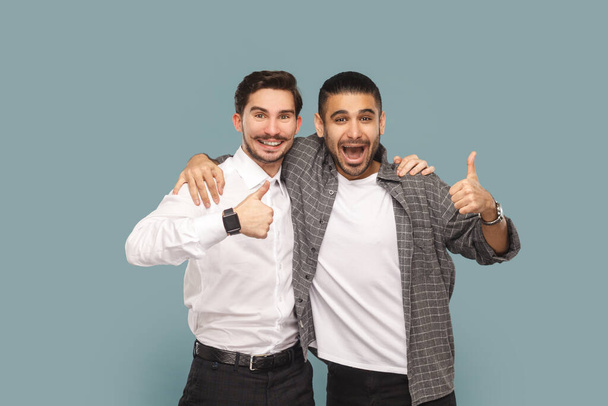 Portrait of two satisfied men friends, standing together and hugging, showing thumbs up, recommend service, good feedback, Indoor studio shot isolated on light blue background. - Foto, Bild