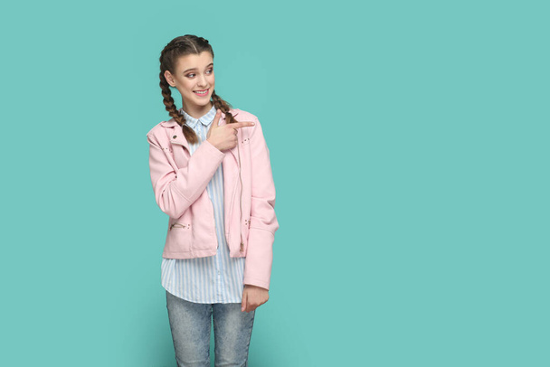 Portrait of smiling positive happy teenager girl with braids wearing pink jacket pointing aside at advertisement area, space for promotion. Indoor studio shot isolated on green background. - Foto, Imagem