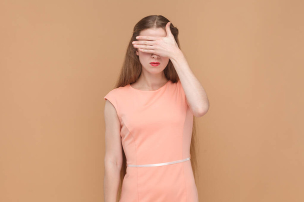Portrait of serious sad woman with long hair standing and covering her eyes with her palms, doesn't want to see something shame, wearing elegant dress. Indoor studio shot isolated on brown background. - 写真・画像