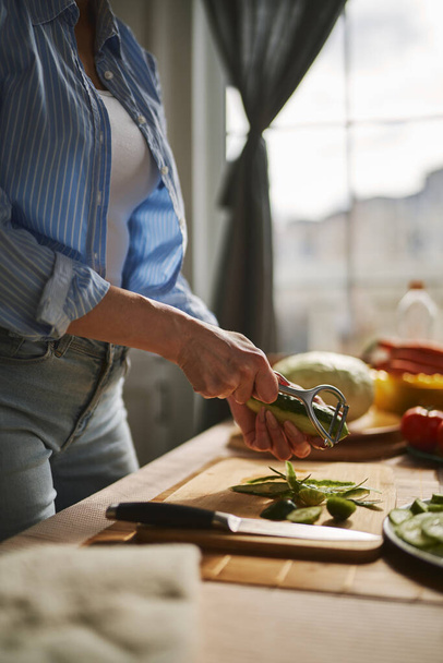 Woman peeling a cucumber for lunch. Housewife using a peeler tool to peel off a vegetables skin - Foto, afbeelding