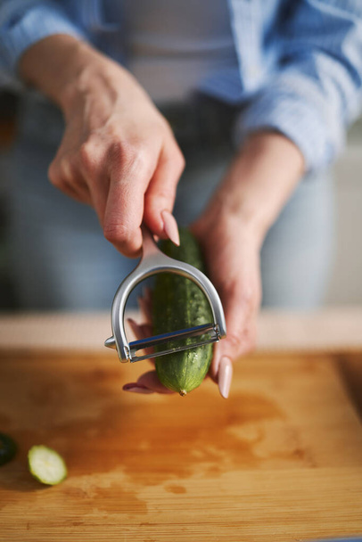 Woman peeling off a cucumber with a peeler tool in close up. Female person preparing a fresh vegetable salad at home - Foto, afbeelding
