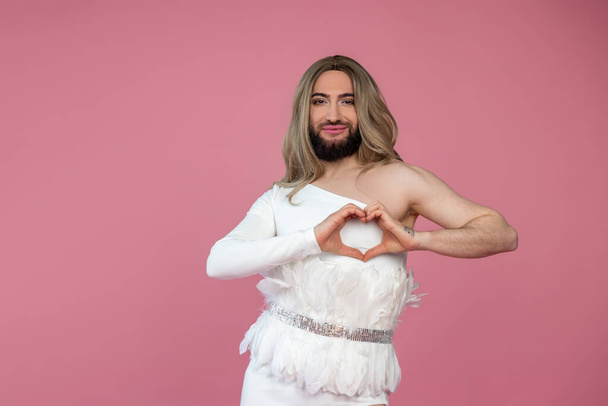 Smiling cheerful transgender wearing blonde wig and white dress making heart shape with hands, expressing love, posing isolated over pink background - Фото, изображение