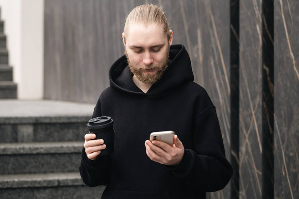 Серйозна молода бородата використовує смартфон і п'є каву. Багаторазовий екологічно чистий сталий зручний бамбуковий стакан нульових відходів, без пластикової концепції
. - Фото, зображення