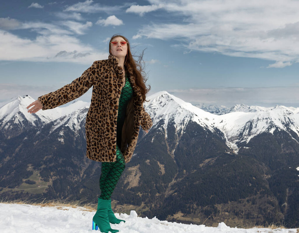 Young woman posing against the backdrop of mountains in a green jumpsuit and a yellow fur coat, with her arms open, Austria. High quality photo - Foto, imagen