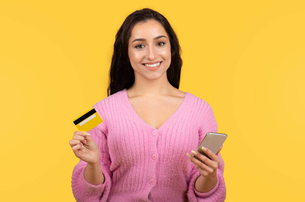 Vrolijke jonge Kaukasische dame in casual met behulp van creditcard en telefoon voor online winkelen geïsoleerd op gele achtergrond, studio. App en gadget voor controle van financiën, bankieren, verkoop, reclame en aanbod - Foto, afbeelding