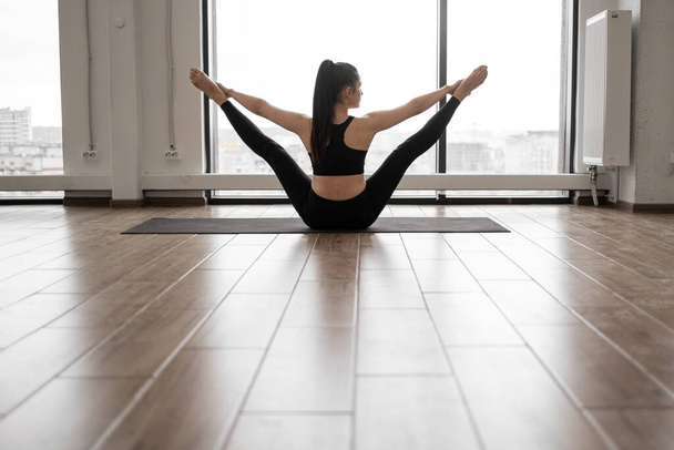 Fitness caucasian woman with dark hair tied in knot flexing body while sitting in leg-splits on yoga mat. Sport center with panoramic windows. Concept of people, sport and active lifestyles. - 写真・画像