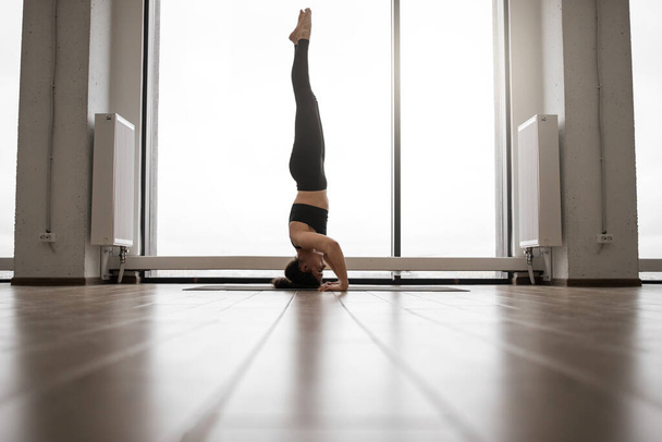 Fit and slender lady doing yoga exercises in black tight leggings and top in front of french windows. Sporty brunette staying in headstand pose at bright commodious studio. Training indoors concept. - Photo, Image