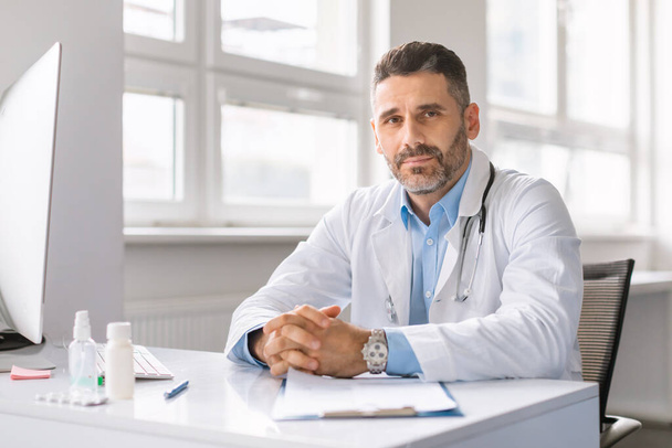 Portrait of confident male doctor in workwear working at modern clinic, sitting at workdesk with computer and looking at camera, copy space - 写真・画像