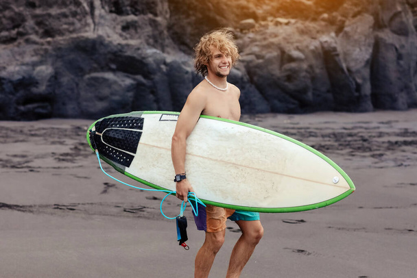 Adatto giovane surfista uomo con i capelli biondi ricci con tavola da surf passa dall'oceano divertendosi facendo sport acquatici estremi, surf. Viaggio e stile di vita sano concetto. Destinazione di viaggio sportiva - Foto, immagini