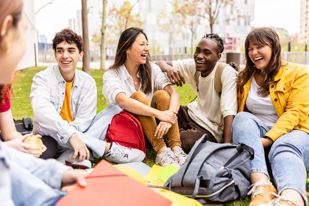 Nuori ryhmä monirotuisia opiskelijoita nauraa ja pitää hauskaa yhdessä istuu nurmikolla yliopiston kampuksella. Teini luokkatoverit sosiaalinen kokoontuminen rentouttava ulkona. - Valokuva, kuva