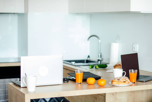 Moderni keittiö, jossa ei ole ihmisiä, joilla on puinen työtaso, jota käytetään kotitoimistona, siinä on kaksi kannettavaa tietokonetta, appelsiineja, kahvikupit ja lasit mehulla. Pääkonttori.  - Valokuva, kuva