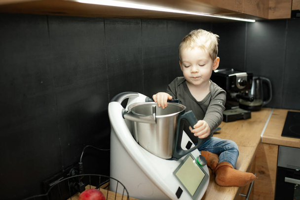 Portrait of concentrated little boy baby toddler with fair hair sitting on table near kneader cooking machine in black kitchen at home, holding metal bowl, looking inside. Childhood, baking, pastry. - Photo, image