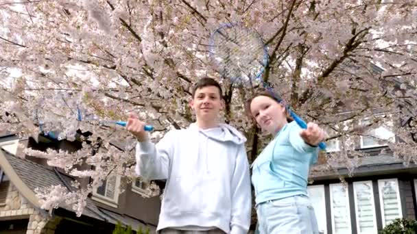 カナダの公園で一緒に立っているバドミントンラケットを持つカップルバンクーバーの子供たちは、ラケットで木の手を開花させることを背景に立っています  - 映像、動画