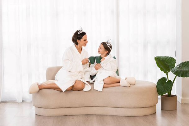 Празднование Дня матери. Милая маленькая девочка и ее мама отдыхают в халатах дома, счастливая молодая женщина и дочь носят игрушечные короны, сидят на диване и пьют горячий шоколад, копируют пространство - Фото, изображение