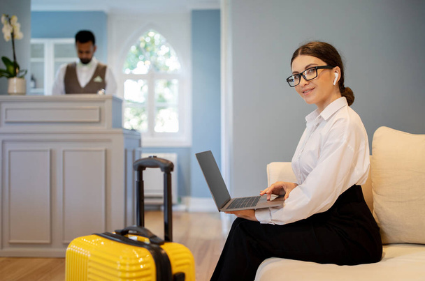 Businesswoman Using Laptop Wearing Wireless Earphones Smiling To Camera Sitting At Hotel Reception. Lady Booking Travel Accomodation And Working Online Posing With Suitcase. Selective Focus - Photo, Image