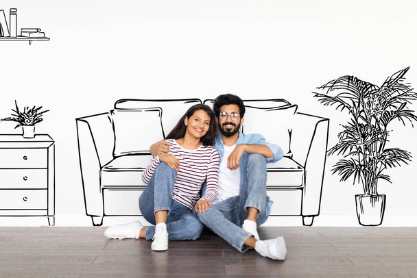 Happy cheerful beautiful young eastern spouses bearded guy wearing eyeglasses and long-haired woman sitting on floor, living room interior drawn on white wall behind, moving to new house, collage - Photo, Image